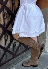 Hellbraune Wildleder-Stiefel bis zum Knie mit dekorativen Nähten, einer spitzen Zehenpartie und einem gestapelten Absatz, kombiniert mit einem weißen Kleid mit Spitze.