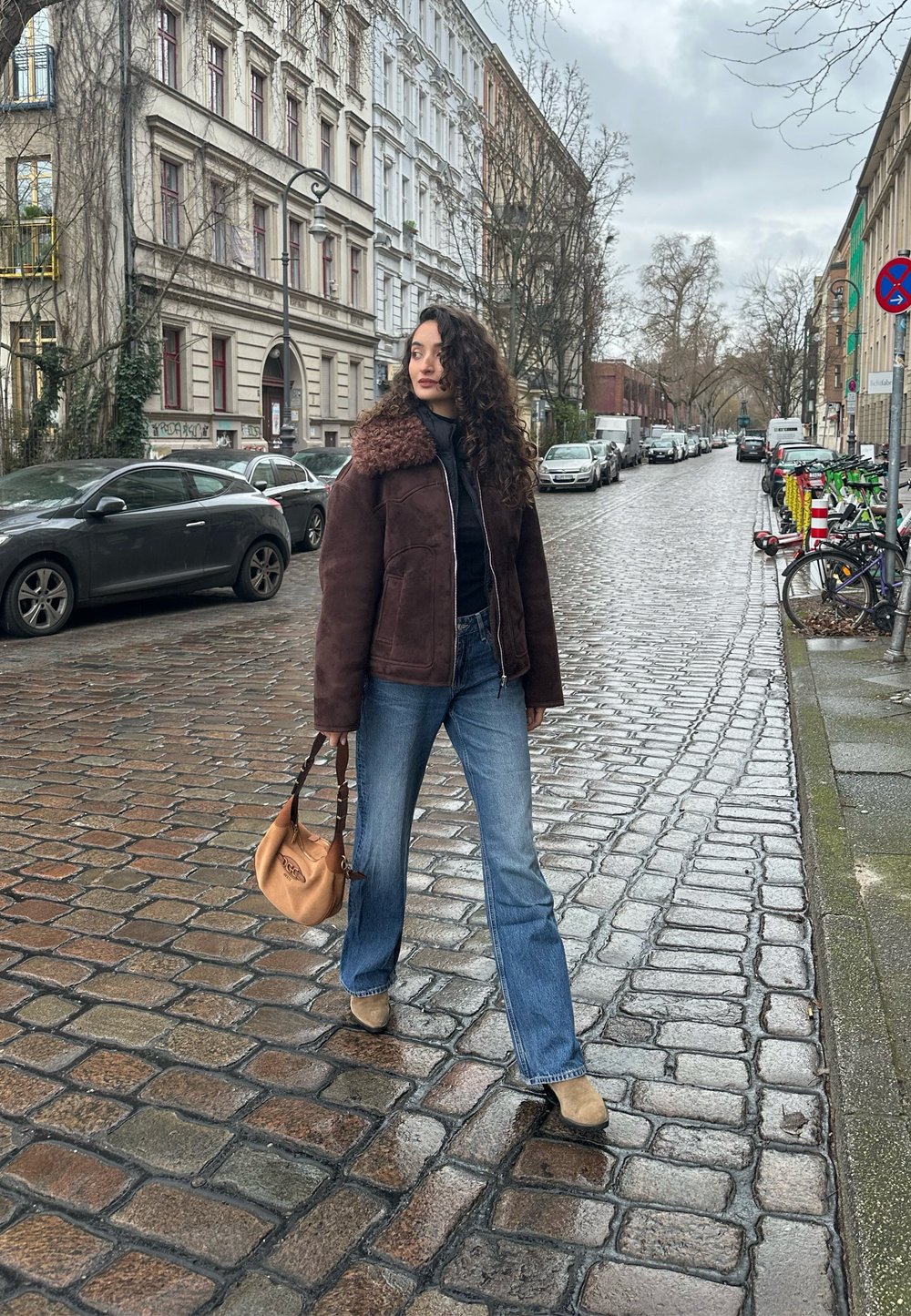Casaco de shearling castanho com gola de pelo, blusa preta, jeans azul boca de sino e botas bege. A carregar uma mala castanha. Fundo de rua de paralelepípedos.