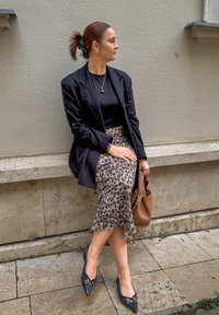 Μαύρο σακάκι πάνω από μια μαύρη μπλούζα, μίντι φούστα με λεοπάρ μοτίβο, μυτερές μαύρες γόβες, κρατώντας μια καφέ τσάντα, καθισμένη σε τοίχο με υφή.