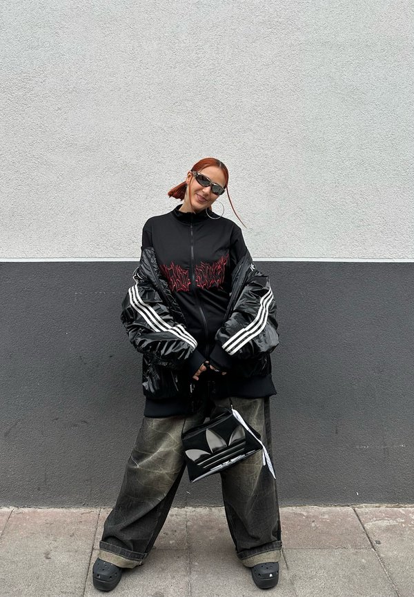 Black puffer jacket with red graphic, black zip-up top, loose grey jeans, black bag with logo, and black clogs; sunglasses complete the look.