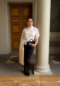 Hvid top med draperet frynse, sort nederdel og sort håndtaske. Omgivelserne inkluderer en trædør og marmorudsmykninger.