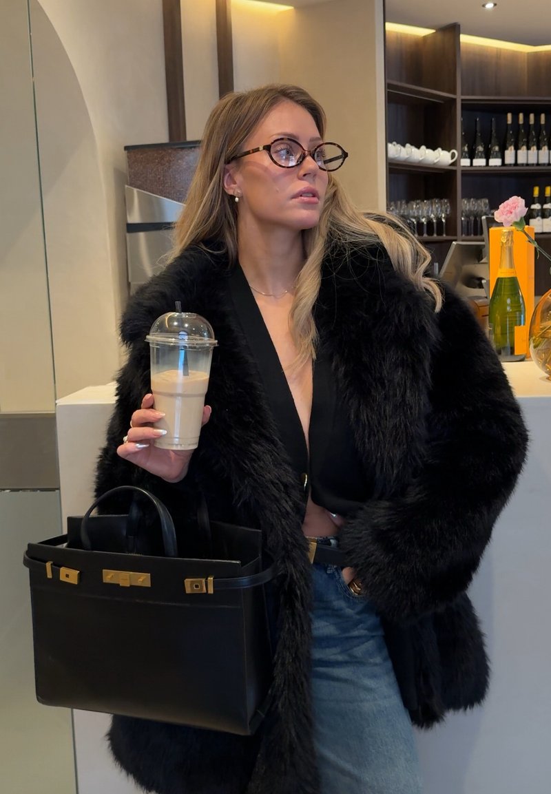 Femme en manteau de fourrure noir et lunettes, tenant un café glacé et un sac à main noir, debout dans un café moderne avec des étagères de bouteilles de vin et de verres.