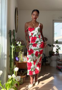 Květinové šaty s červenými hibisky a zelenými listy na bílém pozadí. Špagetová ramínka, přiléhavý střih a délka po kolena.
