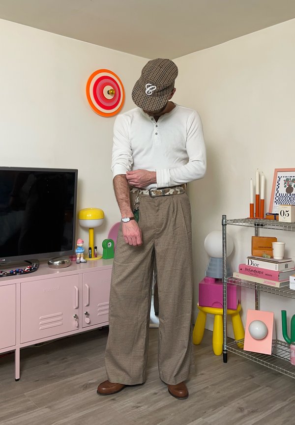 Muž v kostkované čepici, bílém dlouhém rukávu a širokých kostkovaných kalhotách si podhrnuje rukáv v moderním pokoji s barevnou výzdobou.