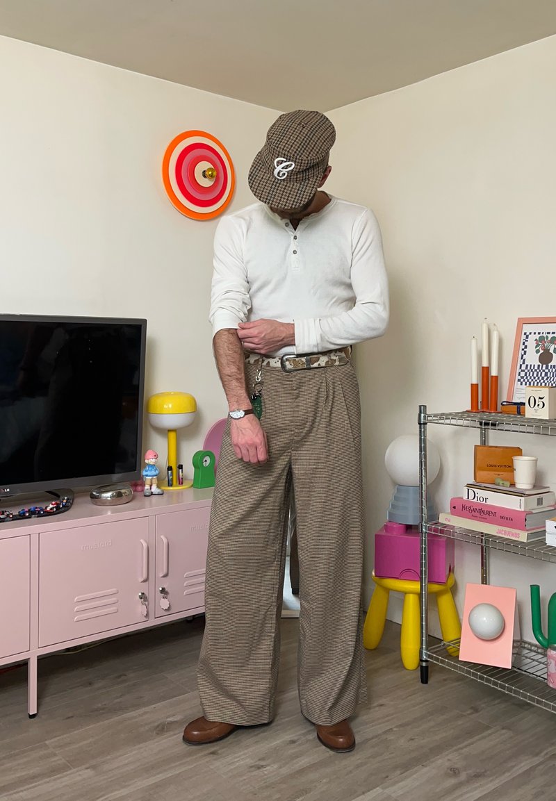 Homme portant une casquette à carreaux, une chemise blanche à manches longues et un pantalon large à carreaux en train de remonter sa manche dans une pièce contemporaine décorée de couleurs vives.