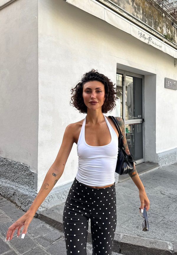 Mujer con el pelo rizado que lleva un top halter blanco y pantalones negros con lunares, sosteniendo gafas de sol y un bolso negro de hombro en una calle de la ciudad.