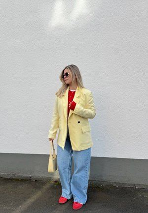Mujer con blazer amarillo, suéter rojo, vaqueros anchos de color azul claro, zapatos rojos, gafas de sol y llevando un bolso beige, de pie contra una pared blanca.
