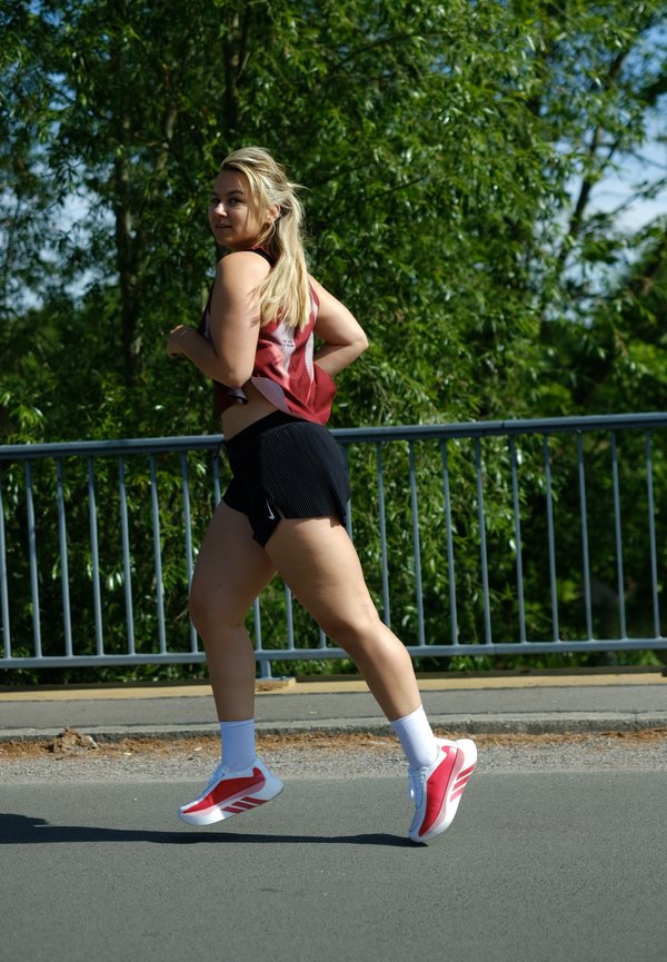 Jeune femme aux cheveux blonds court dehors, portant un short noir, un haut de sport rouge, des chaussettes blanches et des baskets rouges et blanches par une journée ensoleillée.