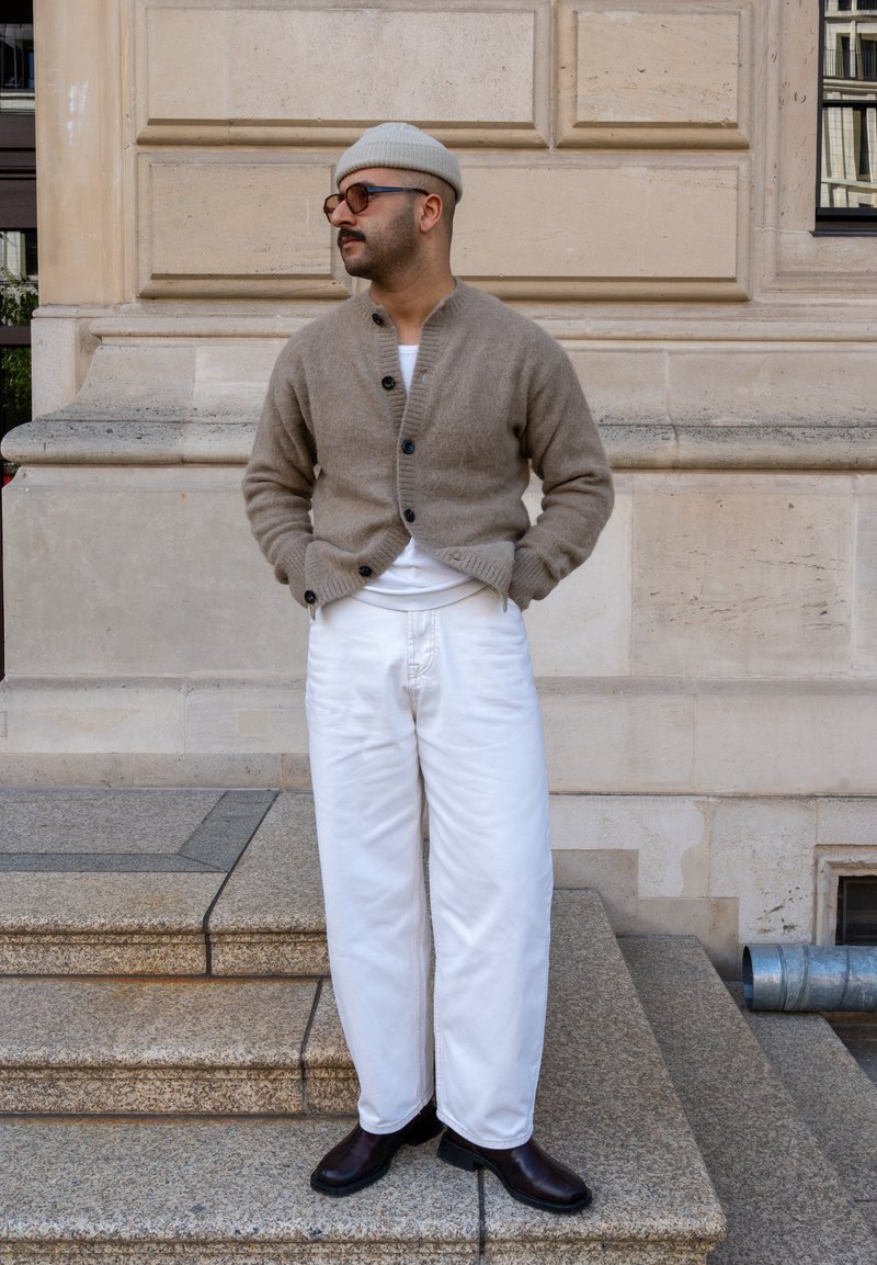 Beiger Wollcardigan mit Knopfverschluss, weisses T-Shirt, lockere weisse Hose und braune Lederschuhe, kombiniert mit einer leichten Mütze und Brille.