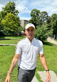 Polo bianco a maniche corte con zip e strisce scure, abbinato a pantaloni a righe grigie. Cappellino bianco con testo ricamato. Sfondo di un parco verde.