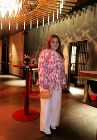 Färgglad mönstrad omlottblus i rosa, grön och vit. Vita vida byxor och en liten orange handväska. Milj ö med trä och modern inredning.