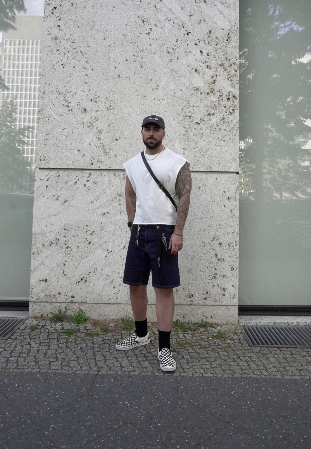 Weißes ärmelloses Shirt, marineblaue Denim-Shorts und schwarz-weiße karierte Sneakers. Enthält eine Umhängetasche und sichtbare Tätowierungen.