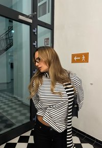 Črtast pulover v črni in beli barvi, ohlapnega kroja, združen s temnimi kavbojkami. Dodatki vključujejo torbo s črtami in okrogla očala. Sodobno notranje okolje v ozadju.