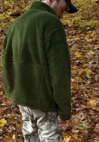Oliwkowa polarowa kurtka z wysokim kołnierzem i teksturowaną powierzchnią, noszona na jasne spodnie w kamuflażu wśród jesiennych liści.
