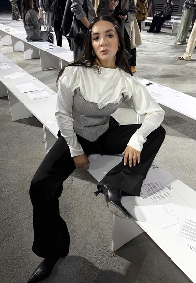 Modelo con una blusa blanca de manga larga con detalles plisados, lleva un corsé gris sobre pantalones negros de campana y botas negras de punta, sentada en un banco.