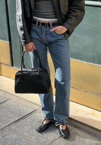 Crna kožna torba, plave traperice, sivi uski top, crne kožne cipele sa vezicama. Modell pozira uz pozadinu u zelenim i zlatnim tonovima.