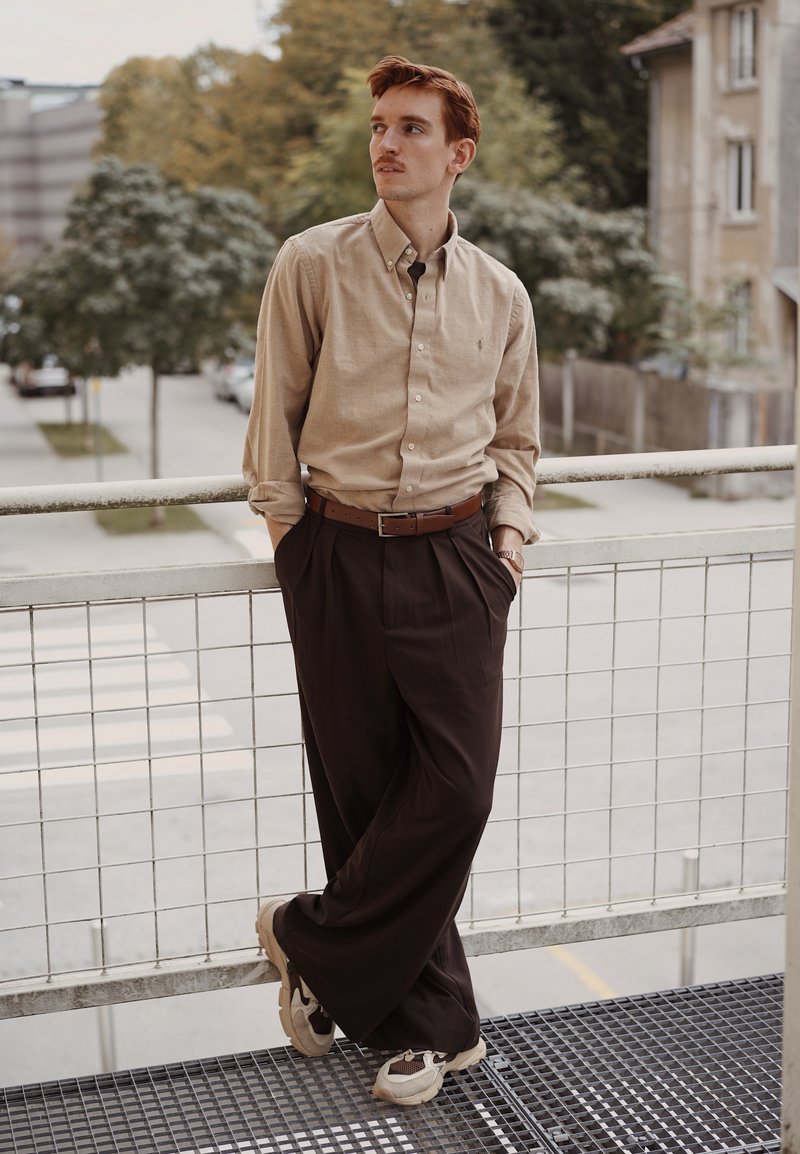 Chemise beige à boutons, ceinture marron, pantalon sombre ample et baskets claires; debout contre une rampe en métal avec un arrière-plan urbain.