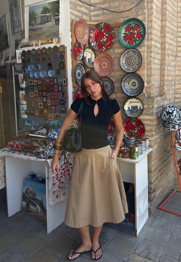 Mujer con blusa negra y falda beige está de pie frente a una pared que exhibe platos decorativos coloridos y una mesa con textiles y cerámicas.
