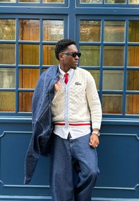 Biały kardigan ze splotem w warkocz z czerwonym i niebieskim wykończeniem, nałożony na białą koszulę, w połączeniu z luźnymi niebieskimi dżinsami i dżinsową kurtką. Noszone okulary przeciwsłoneczne.