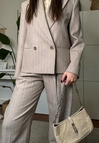 Femme portant un costume beige à fines rayures tenant un sac à main crème avec une chaîne argentée en bandoulière, à l'intérieur, avec des plantes et des étagères en arrière-plan.