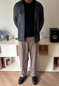 Fekete műbőr ingdzseki, fekete kötött pulóver, világos bézs nadrág és fekete cipő. Világos háttér előtt áll.