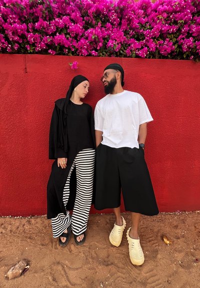 Dos personas están de pie frente a una pared roja. La mujer lleva una blusa negra con un corte y pantalones anchos a rayas. El hombre lleva una camisa blanca y pantalones cortos negros.