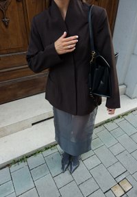 Brązowa marynarka dopasowana z kołnierzem szalowym, w połączeniu z przezroczystą szaroą spódnicą ołówkową i spiczastymi szarymi szpilkami. Czarna torebka o strukturalnym designie.