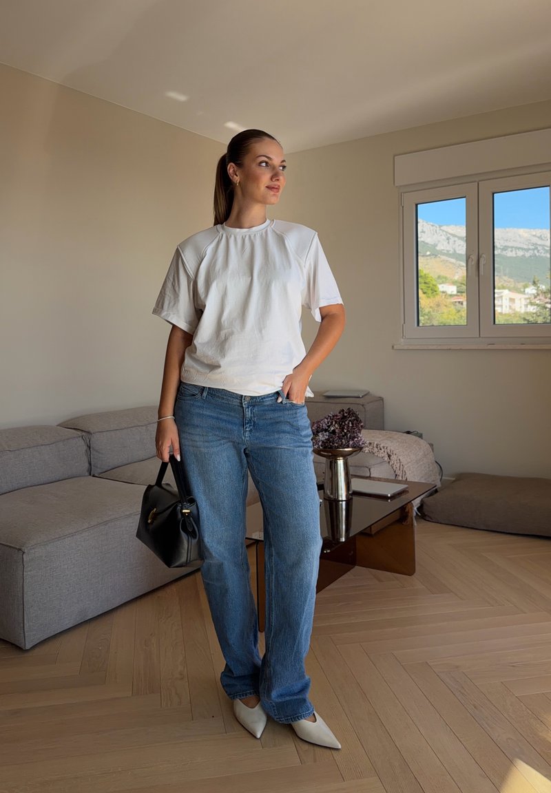 Une femme en T-shirt blanc, en jean bleu et en chaussures plates à bout pointu pose avec un sac à main noir dans un salon. On aperçoit des montagnes par la fenêtre.
