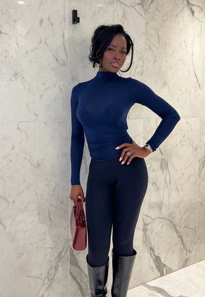 Mujer con cuello alto azul marino, leggings negros y botas hasta la rodilla posa contra una pared de mármol, sosteniendo un bolso burdeos.