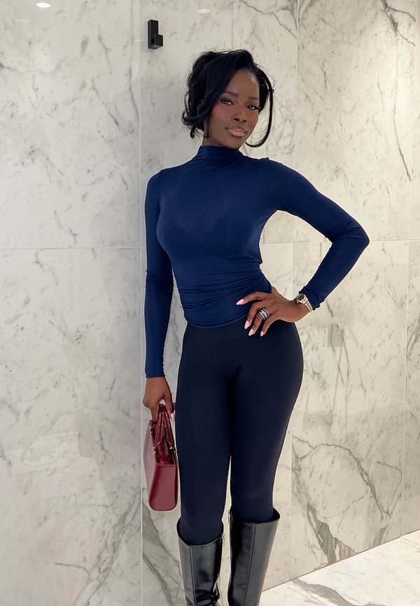 Femme en col roulé bleu marine, leggings noirs et bottes hautes pose contre un mur de marbre, tenant un sac à main bordeaux.