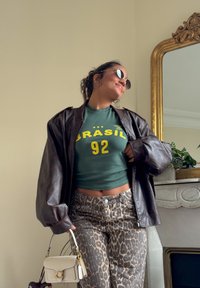 Brown leather oversized jacket, green cropped t-shirt with "BRASÍL" and "92," leopard print high-waisted jeans, and a small cream handbag.