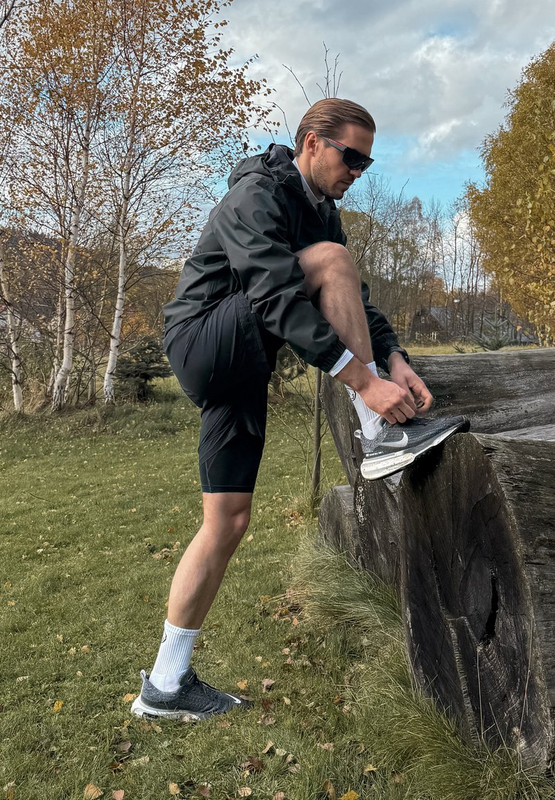 Leggera giacca nera, pantaloni corti neri, calze bianche e scarpe sportive scure. Uomo che allaccia una scarpa su un grande tronco in un'area erbosa con alberi.