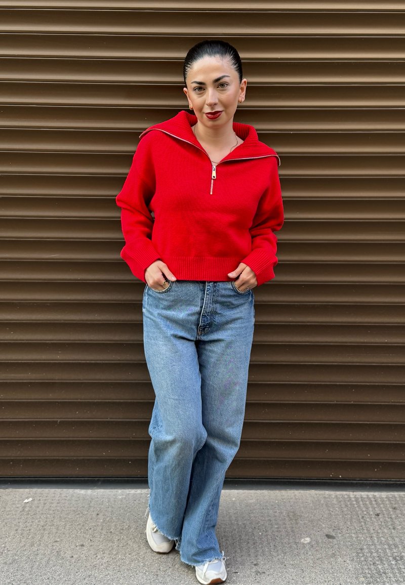 Un pull rouge zippé avec des poignets et un ourlet côtelés, associé à un jean flare bleu clair et des baskets blanches. En arrière-plan, un mur texturé marron.