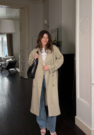 Mujer sonriendo en interior con abrigo beige, camisa blanca con lunares, vaqueros anchos, sandalias blancas y llevando un bolso negro de hombro.