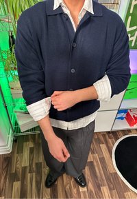 Marineblauwe geribde cardigan met knopen, over een wit gestreept shirt. Grijze broek en zwarte instappers maken de outfit compleet. Schoonmaakgereedschap en dozen op de achtergrond.