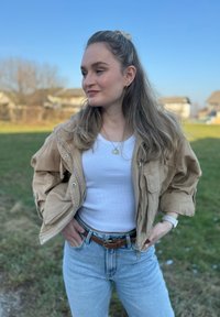 Casaco de fecho éclair castanho claro com dois bolsos frontais sobre uma blusa de alças branca, combinado com jeans de cintura alta azul claro e um cinto castanho.