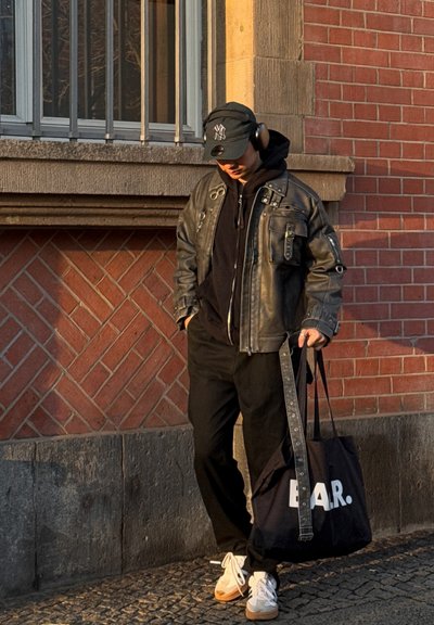 Chaqueta de cuero negra con herrajes plateados, sudadera oscura, pantalones negros, zapatillas blancas y una bolsa de mano azul marino con letras blancas.