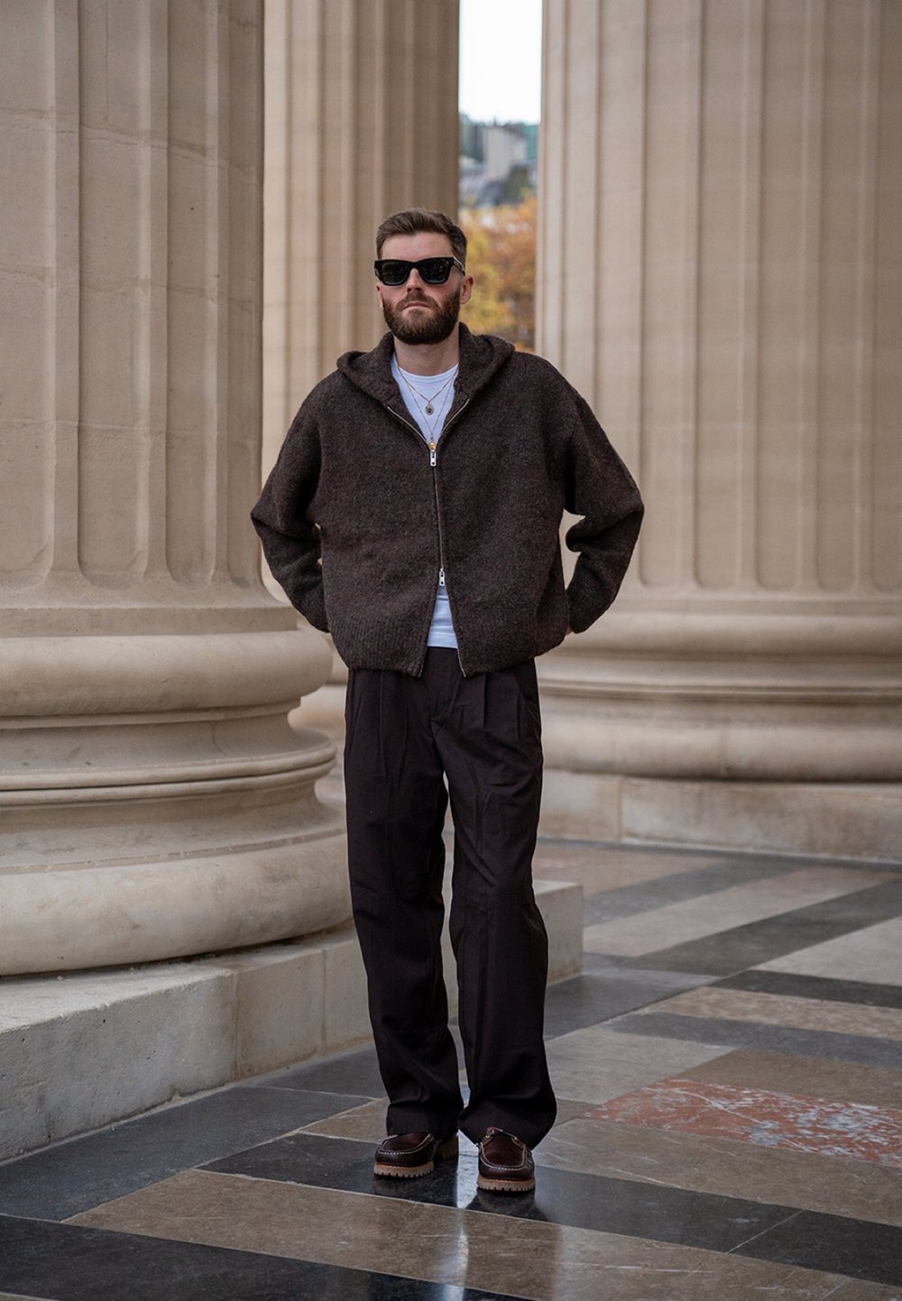 Pull marron à fermeture éclair en maille avec capuche, t-shirt blanc en dessous, pantalon foncé ajusté, et chaussures marron avec une semelle texturée.