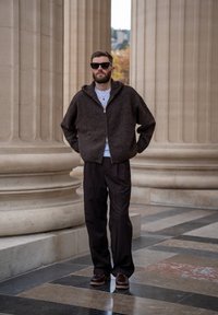 Pull marron à fermeture éclair en maille avec capuche, t-shirt blanc en dessous, pantalon foncé ajusté, et chaussures marron avec une semelle texturée.