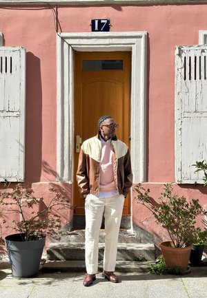 Hombre con chaqueta marrón y beige, pantalones blancos y mocasines, de pie frente a una puerta de madera de una casa rosa con el número 17.