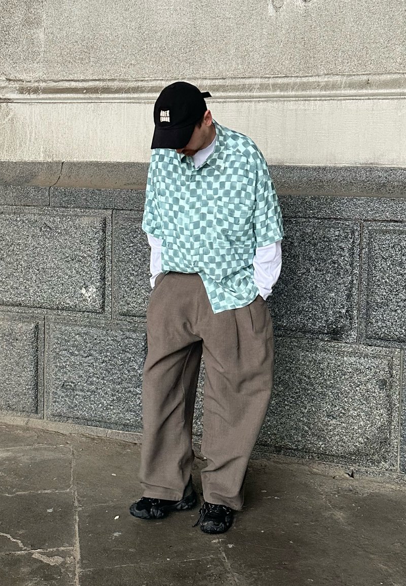 Personne portant une casquette noire, une chemise à manches courtes à carreaux verts par-dessus un t-shirt blanc à manches longues, un pantalon marron ample et des chaussures noires, debout près d'un mur en pierre.