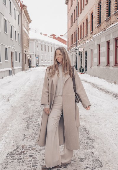 Mujer con abrigo de lana beige y pantalones anchos de pie en una calle adoquinada nevada bordeada de edificios de ladrillo y madera en invierno.