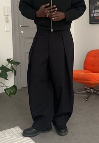 Personne portant un pantalon noir à jambes larges, des chaussures noires et une veste noire zippée, debout à l'intérieur près d'une chaise orange et d'une plante.