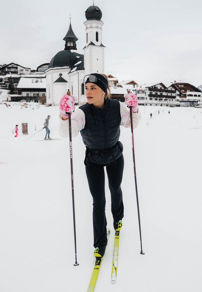 Mujer vestida de blanco y negro esquiando sobre la nieve con guantes rosas y una iglesia con torres con cúpula en el fondo nevado.