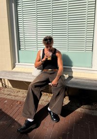 Svart tank top, brune videbukser, sorte sko med hvite sokker, drikker iskald kaffe, sitter på en benk av tre mot et vindu.
