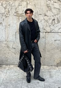 Casaco longo de pele preto, t-shirt preta, jeans escuros e sapatos pretos. A segurá uma bolsa preta com tachas prateadas. Fundo de parede cinza texturizada.