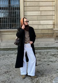 Pikk must mantel pruuni kampsuni, valgete avarad püksid ja beežide saapade üle. Aksessuaarideks on prillid ja pruuni käsikott.