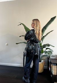 Femme aux longs cheveux blonds portant une veste noire brillante, un pantalon foncé et des chaussures noires, debout à l'intérieur à côté d'une grande plante en pot et d'une étagère.