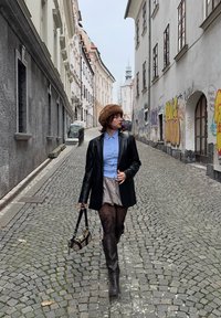 Séta egy köves utcán, fekete bőr blézert, kék inget, mintás szoknyát, barna kalapot és térdig érő barna csizmát viselő személlyel.