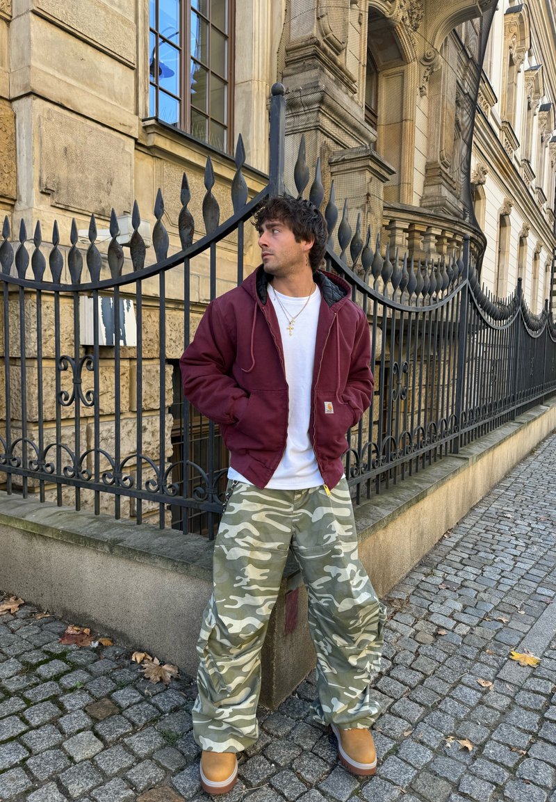 Μπορντό μπουφάν με κουκούλα και μαύρη επένδυση, λευκό μπλουζάκι, πράσινα καμουφλάζ παντελόνια cargo και καφέ υποδήματα μπροστά σε μια πέτρινη πολυκατοικία και σιδερένιο φράκτη.